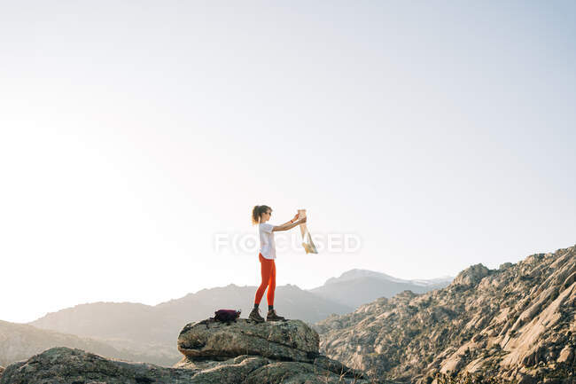Боковой вид на модного молодого туриста в повседневной одежде, запускающего воздушного змея, стоящего на каменном валуне в живописной горной долине под безоблачным голубым небом — стоковое фото