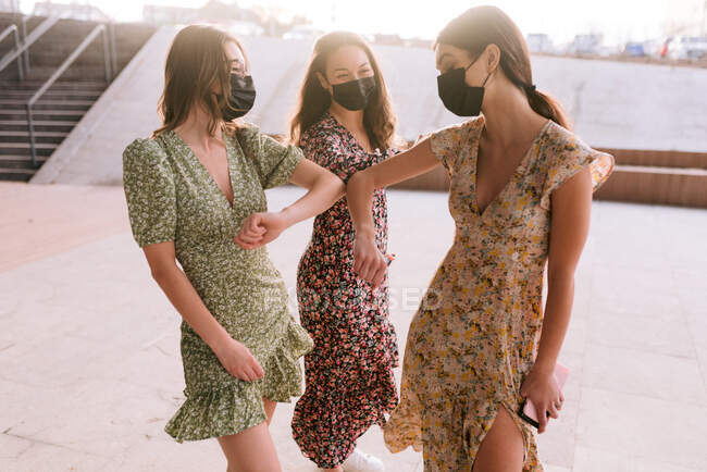 Лучшие подруги в декоративных платьях и масках для лица, касающихся локтей, глядя друг на друга в городе во время пандемии коронавируса — стоковое фото