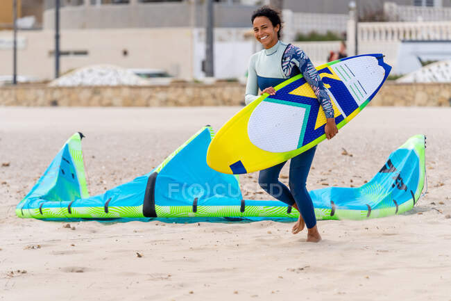 Cheerful ethnic female athlete in wetsuit with kiteboard running on sandy shore against inflatable kite while looking at camera — Stock Photo