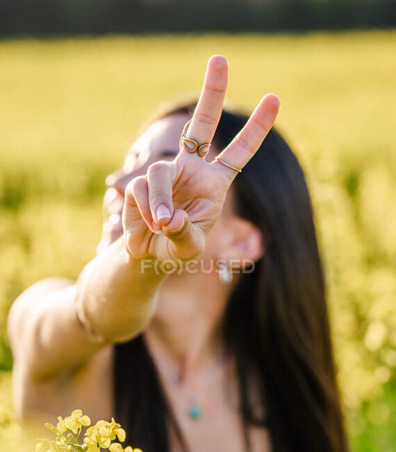 Jovem morena alegre mostrando dois dedos gesto para câmera enquanto desfruta de dia ensolarado no campo de colza florescente — Fotografia de Stock