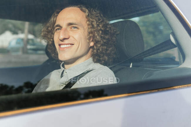 Through window view of smiling young stylish haired male passenger sitting on backseat of modern automobile and fastened with safety belt enjoying trip — Photo de stock