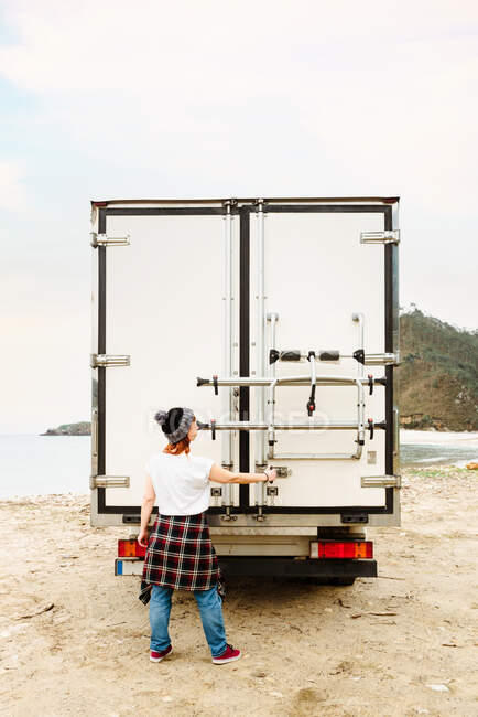 Vista trasera de la puerta de apertura hembra hipster de camión estacionado en la playa cerca del lago durante el viaje de verano - foto de stock