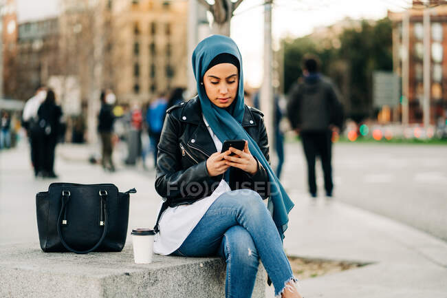 Mujer musulmana en pañuelo para la cabeza sentada en el banco con bebida para llevar y bolso mientras navega por el teléfono móvil en la ciudad - foto de stock