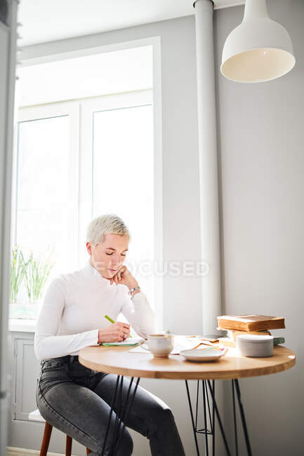 Female astrologist taking notes in notepad at desk with cup of coffee at home in sunlight — Stock Photo