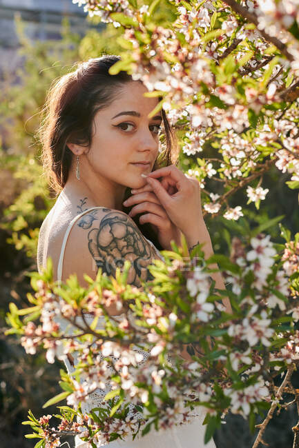 Young female with tattooed arm wearing white dress and standing in flowers of tree looking at camera — Fotografia de Stock