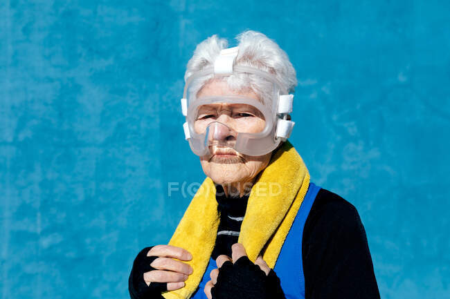 Femme mûre sérieuse en vêtements de sport en protège-tête de boxe avec serviette sur l'épaule debout sur fond bleu et regardant la caméra — Photo de stock