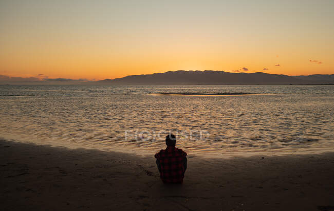 Back view of unrecognizable tourist sitting alone on sandy beach near calm sea and contemplating picturesque sunset — Stock Photo