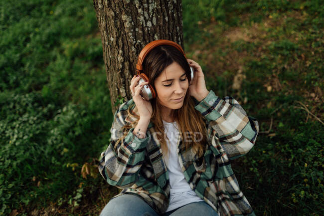 Молодая женщина с татуировкой в клетчатой рубашке слушает музыку из наушников на газоне летом — стоковое фото