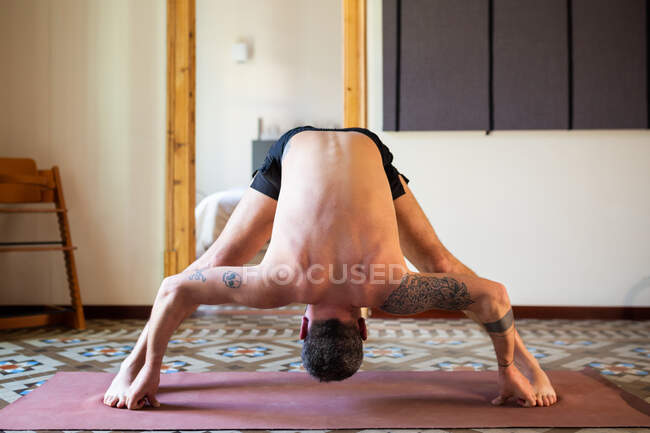 Flexibler anonymer Mann mit nacktem Oberkörper steht in Prasarita Padottanasana, während er zu Hause Yoga praktiziert und seinen Körper dehnt — Stockfoto