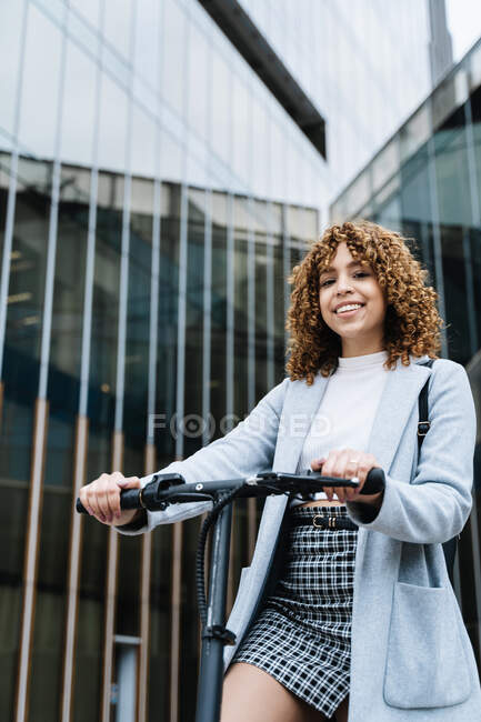 Молода афроамериканська жінка в стильному одязі стоїть з скутером на вулиці і дивиться на камеру з посмішкою. — стокове фото