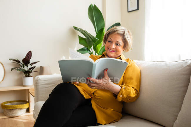 Contenuto Donna di mezza età che si rilassa sul divano e legge una storia interessante mentre si gode il weekend a casa — Foto stock