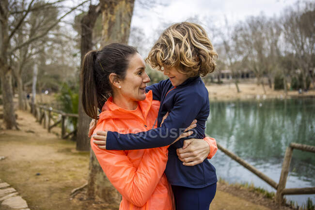Charming boy embracing smiling mother while sitting and looking at each other against water in daylight — Fotografia de Stock