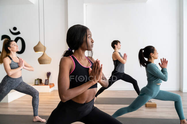 Vista lateral de mujer afroamericana en compañía de diversas personas que practican yoga en Warrior posan en estudio - foto de stock