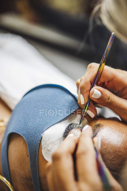 Crop unrecognizable cosmetologist with tweezers applying fake eyelashes for extension on eye of ethnic client with face protective mask in salon during coronavirus pandemic — Stock Photo