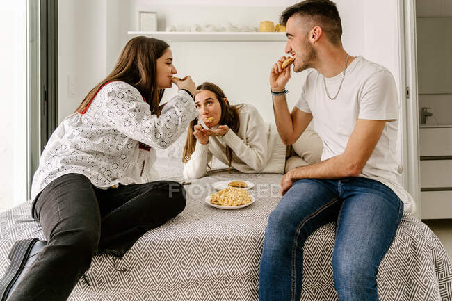 Cheerful young ethnic male and female friends in casual clothes relaxing on bed and eating yummy pastries during weekend at home — Stock Photo
