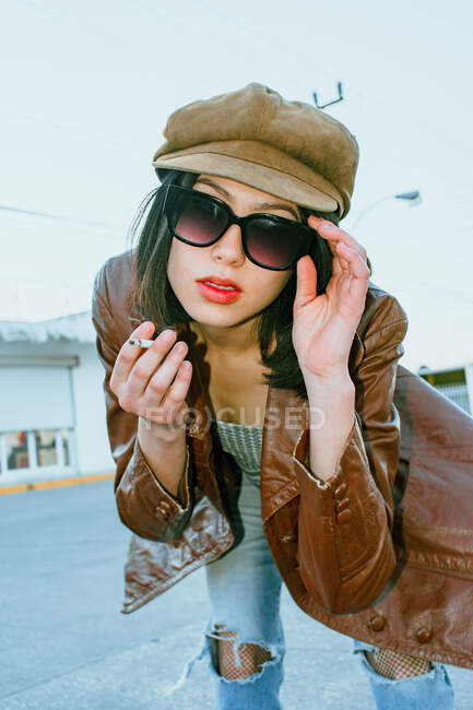 Ethnic female in trendy wear with red lips and cigarette leaning forward on urban road — Stock Photo