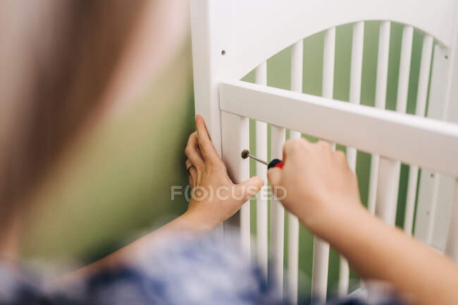 Crop woman with screwdriver leaning forward while mounting crib at home on sunny day — Stock Photo