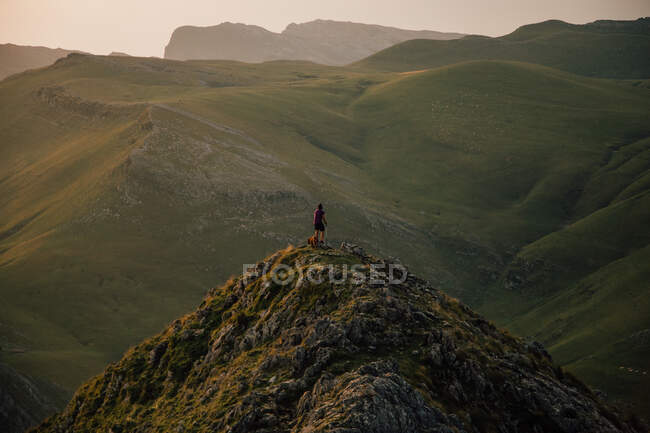 Distant tourist with dog on high peak of mountain range with slopes in green grass under clouds Txindoki, Spain — Stock Photo