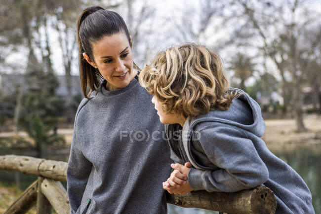 Side view of cheerful mom with boy in casual apparel on wooden fence contemplating nature while looking away — Photo de stock
