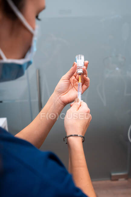 Vista lateral del veterinario femenino irreconocible recortado en tela mascarilla tomando medicamento con inyector del frasco - foto de stock