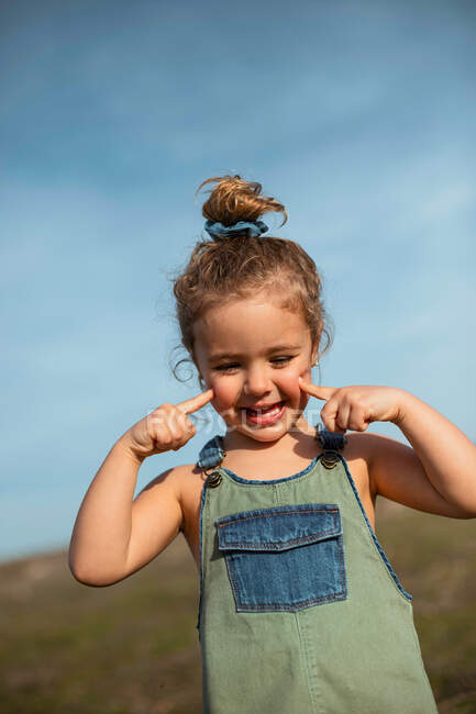 Entzückendes kleines Mädchen in Overalls, das mit den Fingern auf der Wiese steht und nach unten schaut — Stockfoto