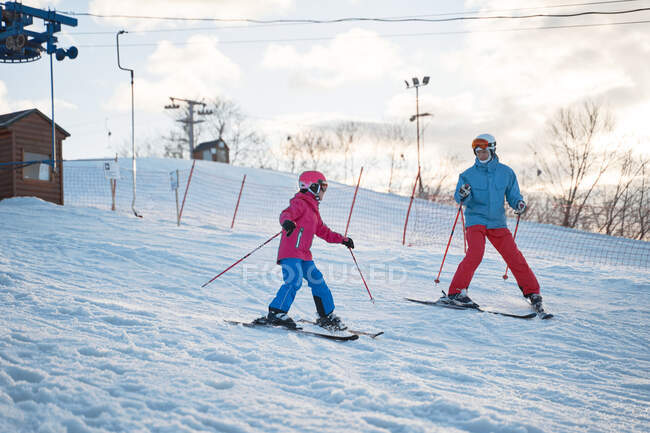 Полнотелый родитель в теплой спортивной одежде и шлеме учит маленького ребенка кататься на лыжах вдоль снежного склона холма на зимнем горнолыжном курорте — стоковое фото