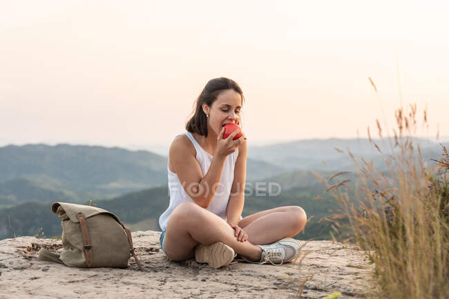 Mujer viajando sentada en la roca y comiendo manzana dulce mientras se relaja durante la caminata en las montañas al atardecer. - foto de stock