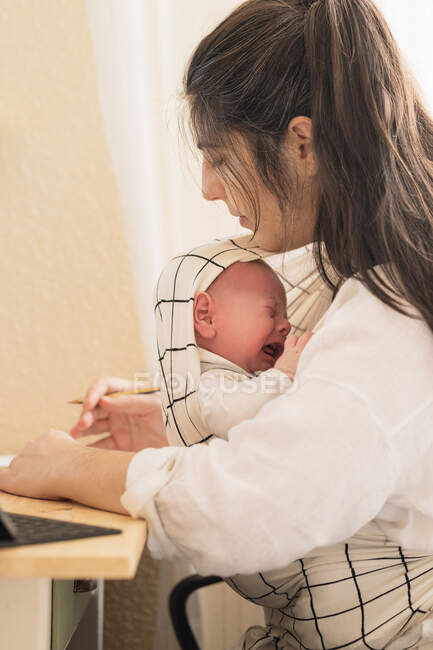 Side view of mother with crying little child in baby carrier sitting at table in house — Stock Photo