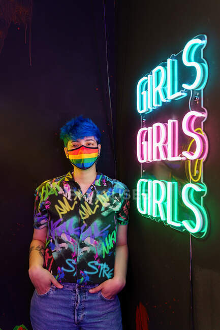 Donna omosessuale in camicia con lettere e maschera tessile guardando la macchina fotografica mentre in piedi con le mani in tasca contro le iscrizioni delle ragazze — Foto stock