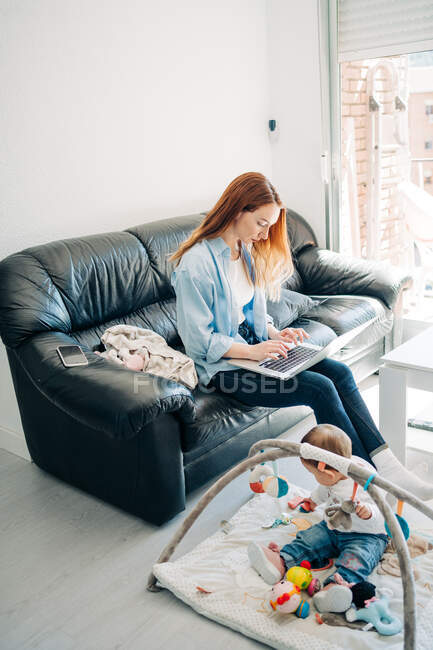 Comprimento total focado jovem mãe em roupa casual netbook de navegação no sofá perto adorável bebê brincando com brinquedos no chão na sala de estar — Fotografia de Stock