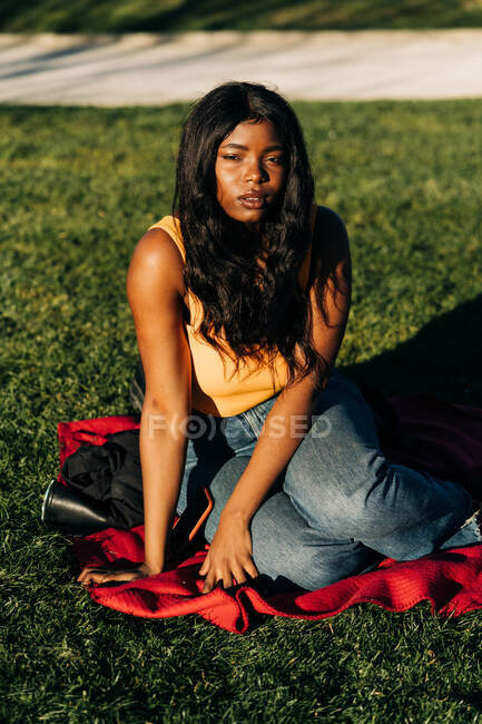 Unbekümmerte Afroamerikanerin im schicken Outfit sitzt auf kariertem Rasen im Park und blickt in die Kamera — Stockfoto