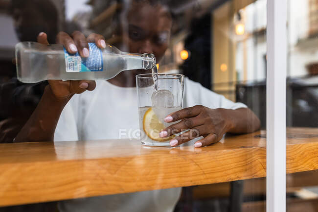 Through glass crop calm African American female in casual outfit pouring cold fresh water from bottle into glass with ice and lemon while sitting at high table in cafeteria — Stock Photo