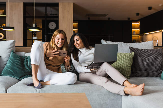 Un par de mujeres homosexuales alegres que se relajan en el sofá y ven videos en teléfonos inteligentes y trabajan en computadoras portátiles mientras se entretienen juntos el fin de semana. - foto de stock