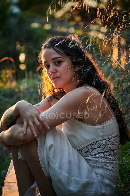 Мрійлива леді з татуйованою рукою в білій сукні і сидить на зеленому газоні в природі — стокове фото