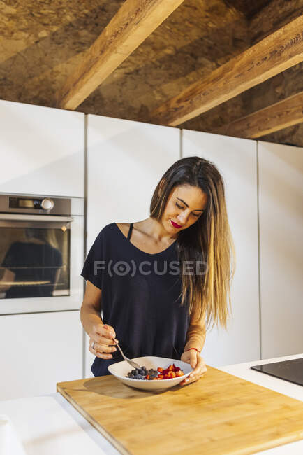 Ehrliche Hündin mit Löffel über Schüssel mit verschiedenen geschnittenen frischen Beeren in Schüssel zu Hause — Stockfoto