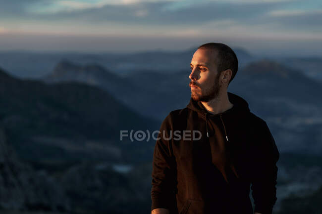 Contemplative bearded male hiker in black hoodie standing on rough mountain summit and observing picturesque highlands in twilight in Seville Spain — Stock Photo