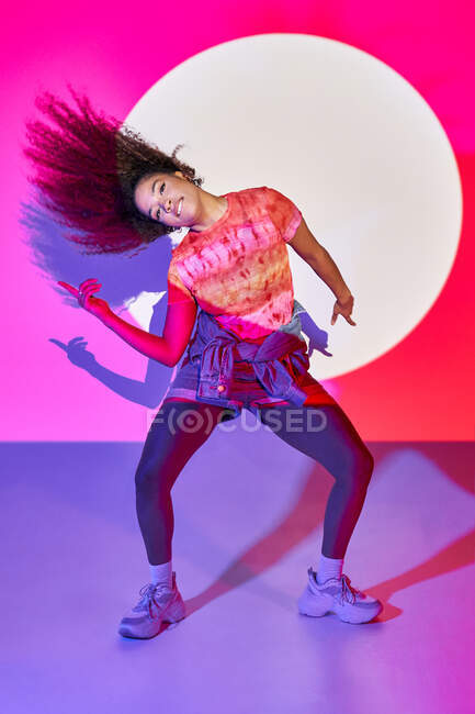 Fit bailarina afroamericana en pantalones cortos bailando mirando a la cámara mientras está de pie en luces de neón en el estudio - foto de stock