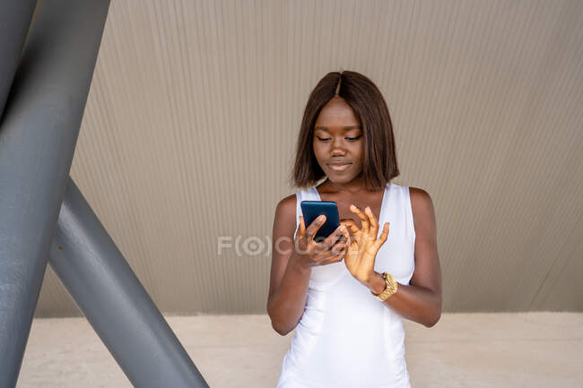 Linda mulher afro-americana em elegante vestido branco que navega telefone celular enquanto está de pé na calçada — Fotografia de Stock