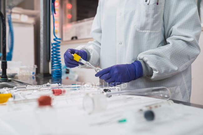 Crop anonymous competent researcher in white robe latex gloves and face mask working in modern laboratory and standing near table with assorted glassware including graduated cylinders syringes and test tubes — Stock Photo