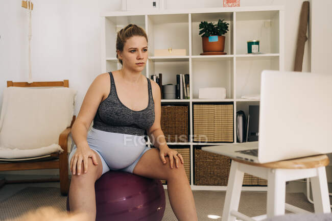 Jovem expectante fêmea sentada na bola de exercício e laptop de navegação na sala de estar — Fotografia de Stock