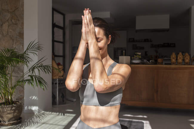 Zufriedene Frau mit Namaste-Händen meditiert im Raum mit Sonnenlicht, während sie zu Hause Yoga praktiziert — Stockfoto
