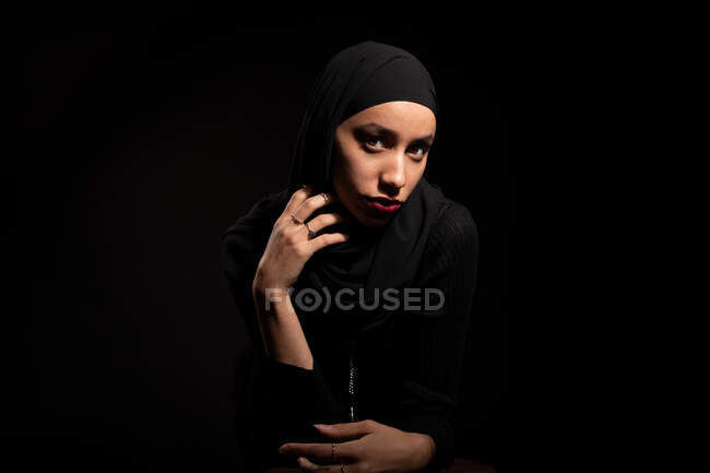 Привлекательная молодая исламская женщина в черной одежде и хиджабе нежно смотрит в камеру на черной студии — стоковое фото