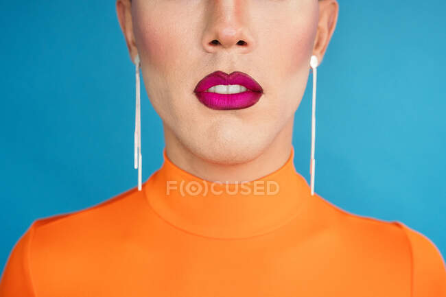 Crop slim female with bright makeup wearing stylish earrings standing against blue wall in studio — Stock Photo