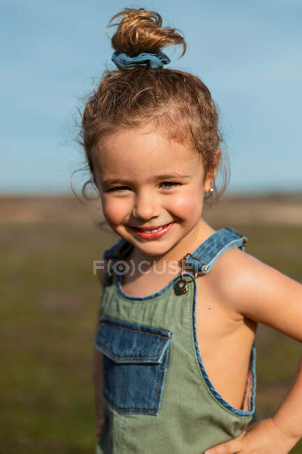 Delighted adorable little girl in overalls standing with hands on waist in meadow and looking at camera — Stock Photo