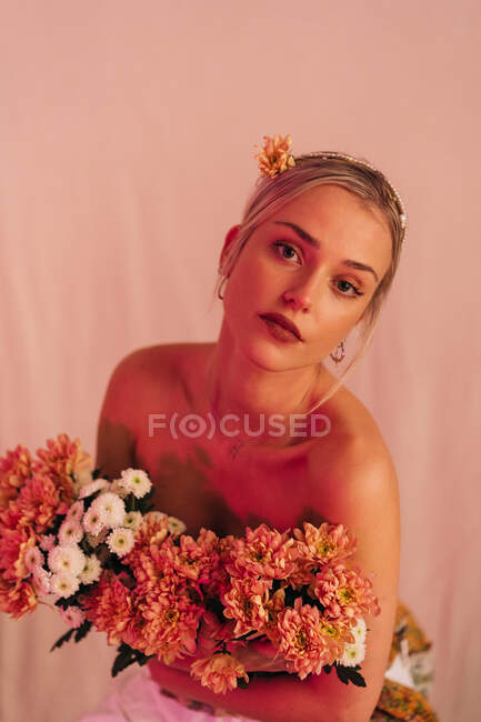 Charming romantic young bare shouldered female with bunch of fresh blooming flowers standing against beige background illuminated with neon light — Stock Photo