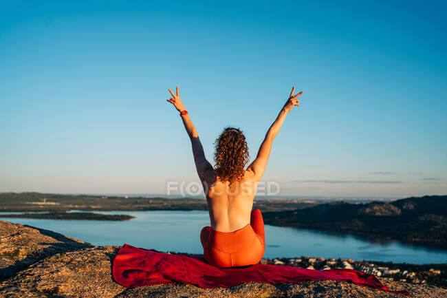 Back view of happy topless female traveler with curly hair in leggings smiling and showing two fingers gesture with raised arms sitting on rocky cliff above coastal town — Stock Photo