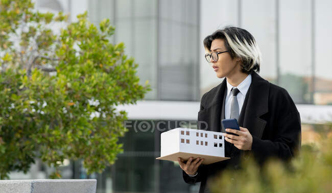 Молодой этнический мужчина исполнительный в очках с макетом дома и сотовым телефоном глядя в город — стоковое фото