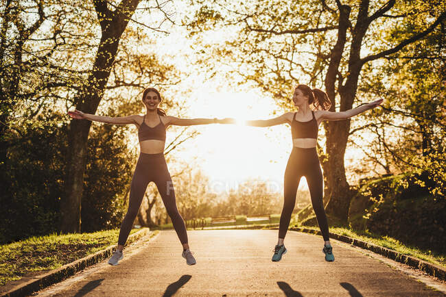 Alegre atlética corredores femeninos de pie en el camino en el parque y calentando los brazos durante el entrenamiento al atardecer - foto de stock