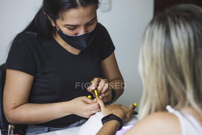 Crop unrecognizable ethnic female master in sterile mask applying yellow polish on nails of anonymous client in beauty salon — Fotografia de Stock