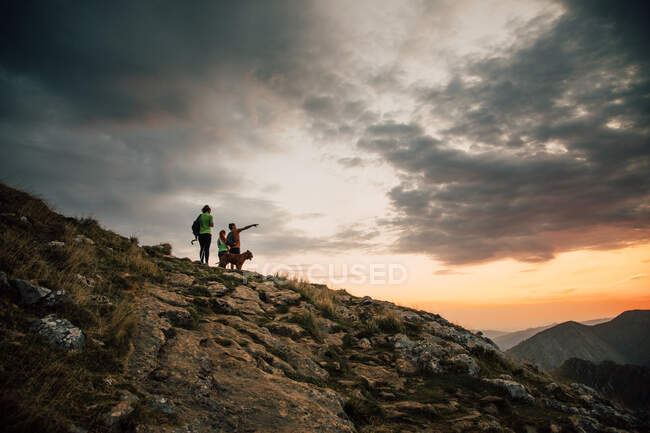 Low angle of group of hikers with dog standing on high peak and observing mountain ridge under sunset sky in clouds — Stock Photo